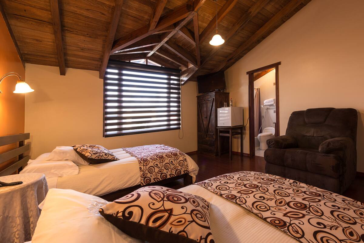 A room with two beds, a wooden ceiling, a chair, and a window with blinds.