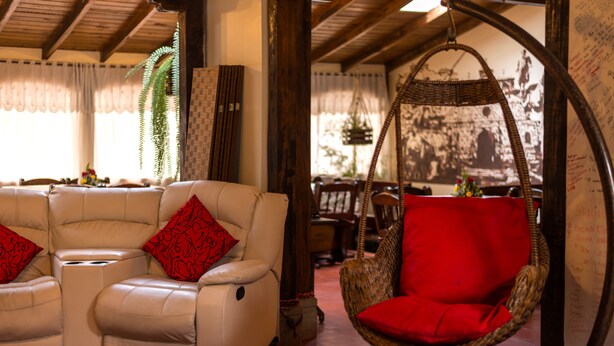 A cozy living area with a white leather sofa and a red cushion, a wicker hanging chair, and a large window with curtains. - Hotel Bella Casona (Riobamba)