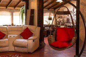 A cozy living area with a white leather sofa and a red cushion, a wicker hanging chair, and a large window with curtains. - Hotel Bella Casona (Riobamba)
