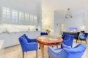 A modern kitchen with white cabinets and a marble countertop, a dining area with a wooden table and blue upholstered chairs, and a living room with a sofa and a TV. - Navy Group - Lion's Castle (Gdansk)
