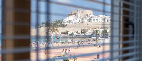 A view through window blinds of a beach area with palm trees, people, and a historic building in the background.