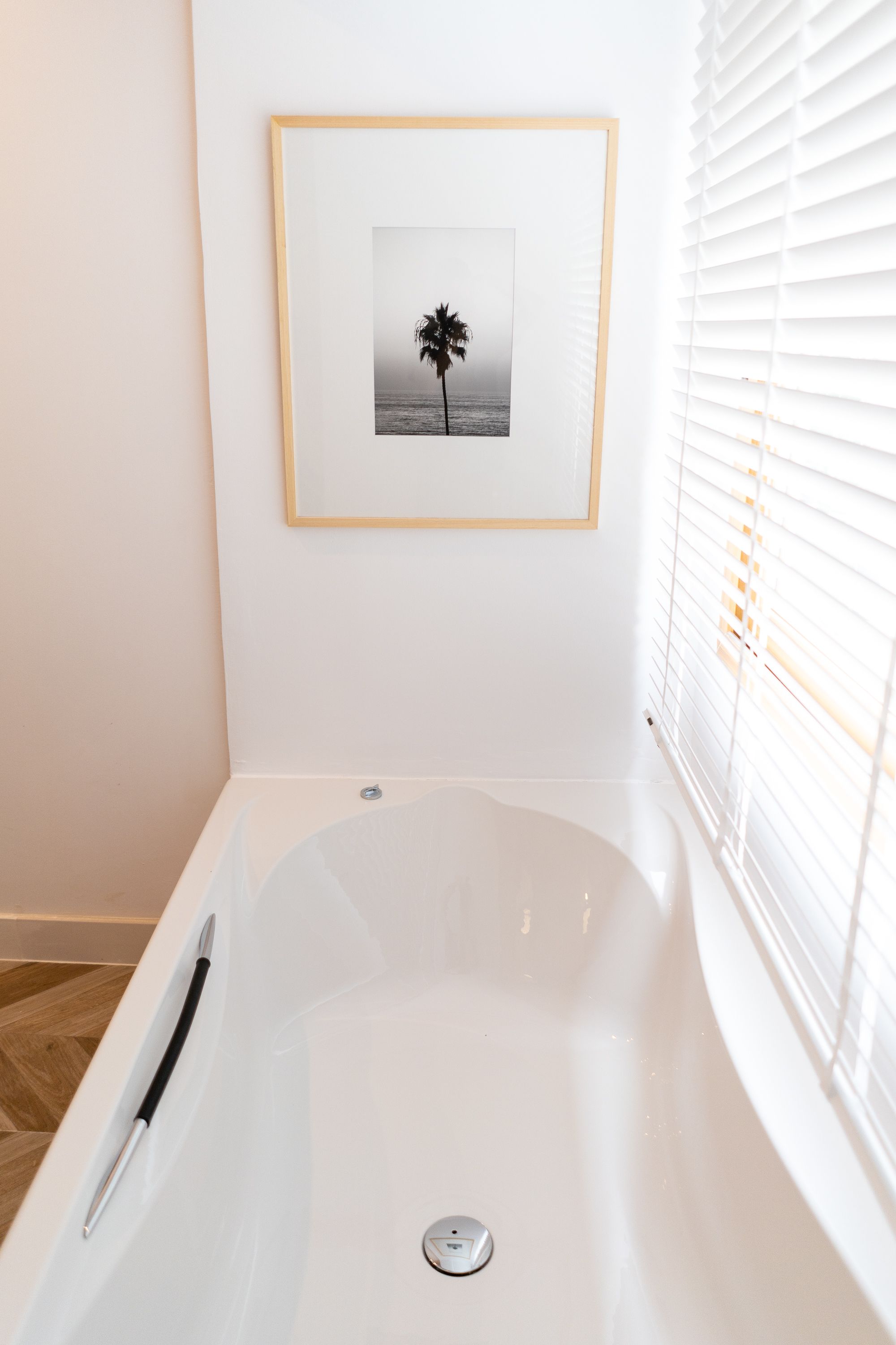A modern bathroom with a white bathtub, a framed picture of a palm tree, and white blinds on the window.