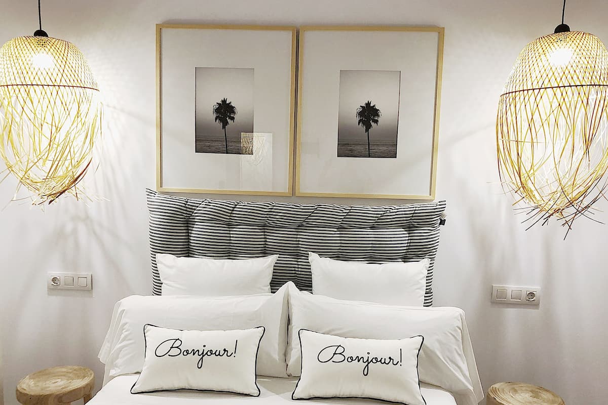 A neatly arranged bedroom with a bed, two pillows with "Bonjour!" text, a striped headboard, and two hanging light fixtures.