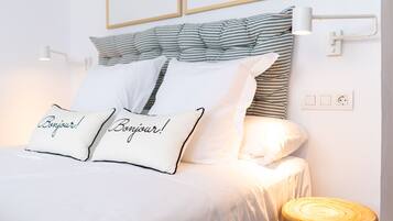 Ein ordentlich bezogenes Bett mit weißer Bettwäsche und Kissen, ein Kopfstück mit Streifenmuster, eine Nachttischlampe und ein hölzerner Hocker.