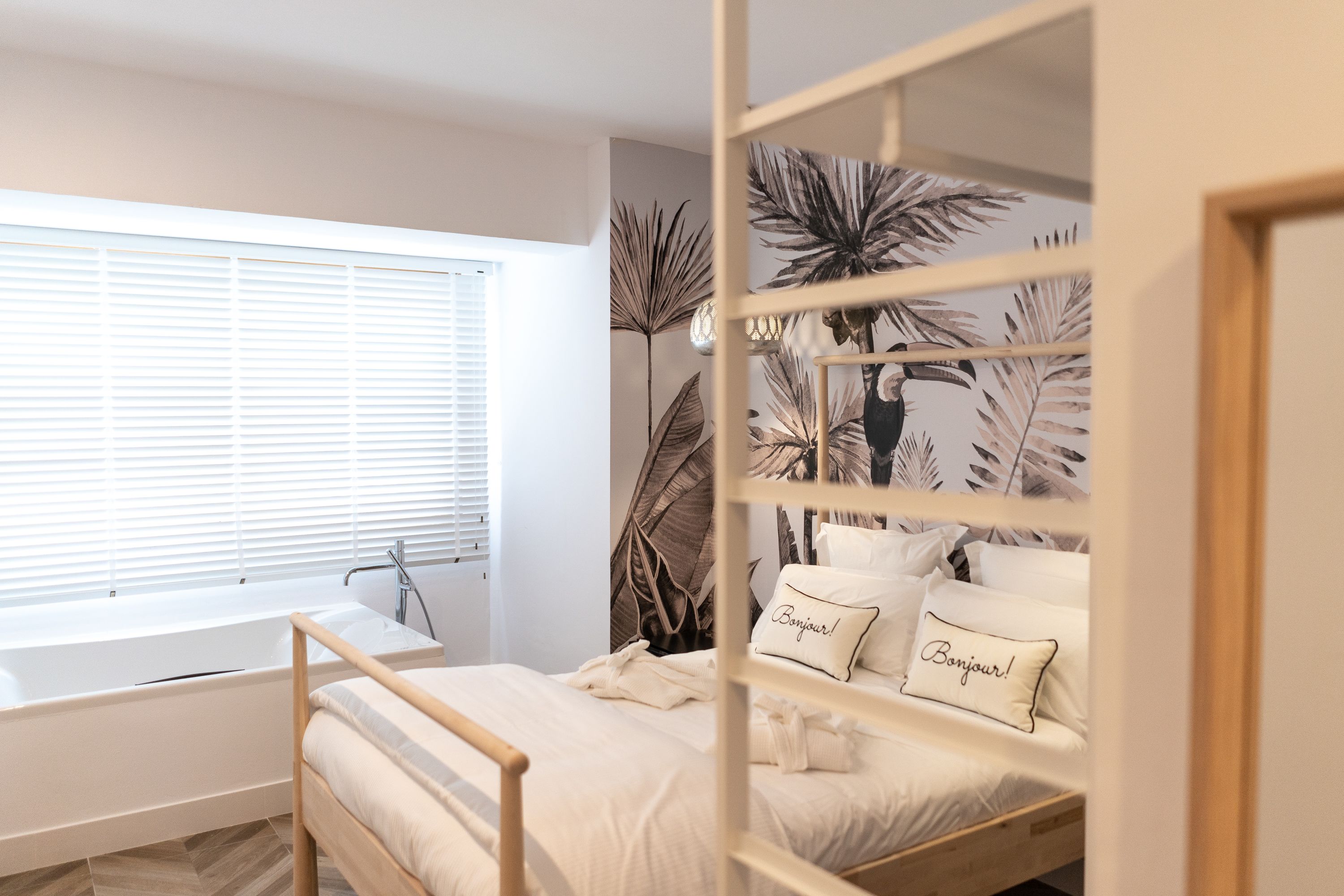 Een slaapkamer met een bed, een raam met zonwering, een badkuip en een muur met een tropisch bladmotief.