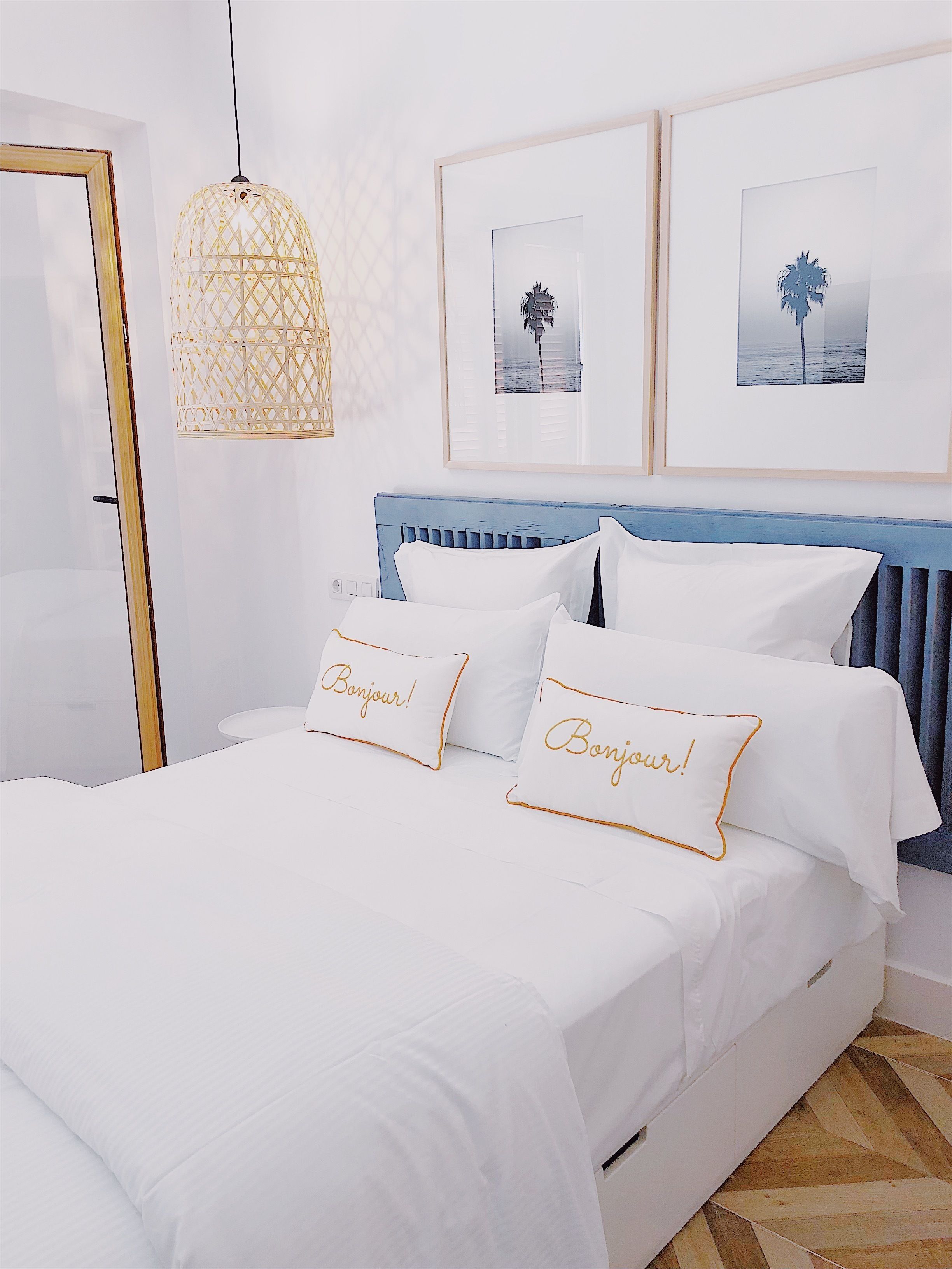 Una cama bien hecha con ropa blanca y dos almohadas que dicen "Bonjour", un cabecero azul y dos cuadros en la pared.
