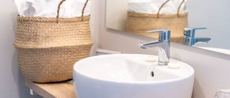 Un baño con un lavamanos blanco, grifo cromado y una cesta tejida en un estante de madera.