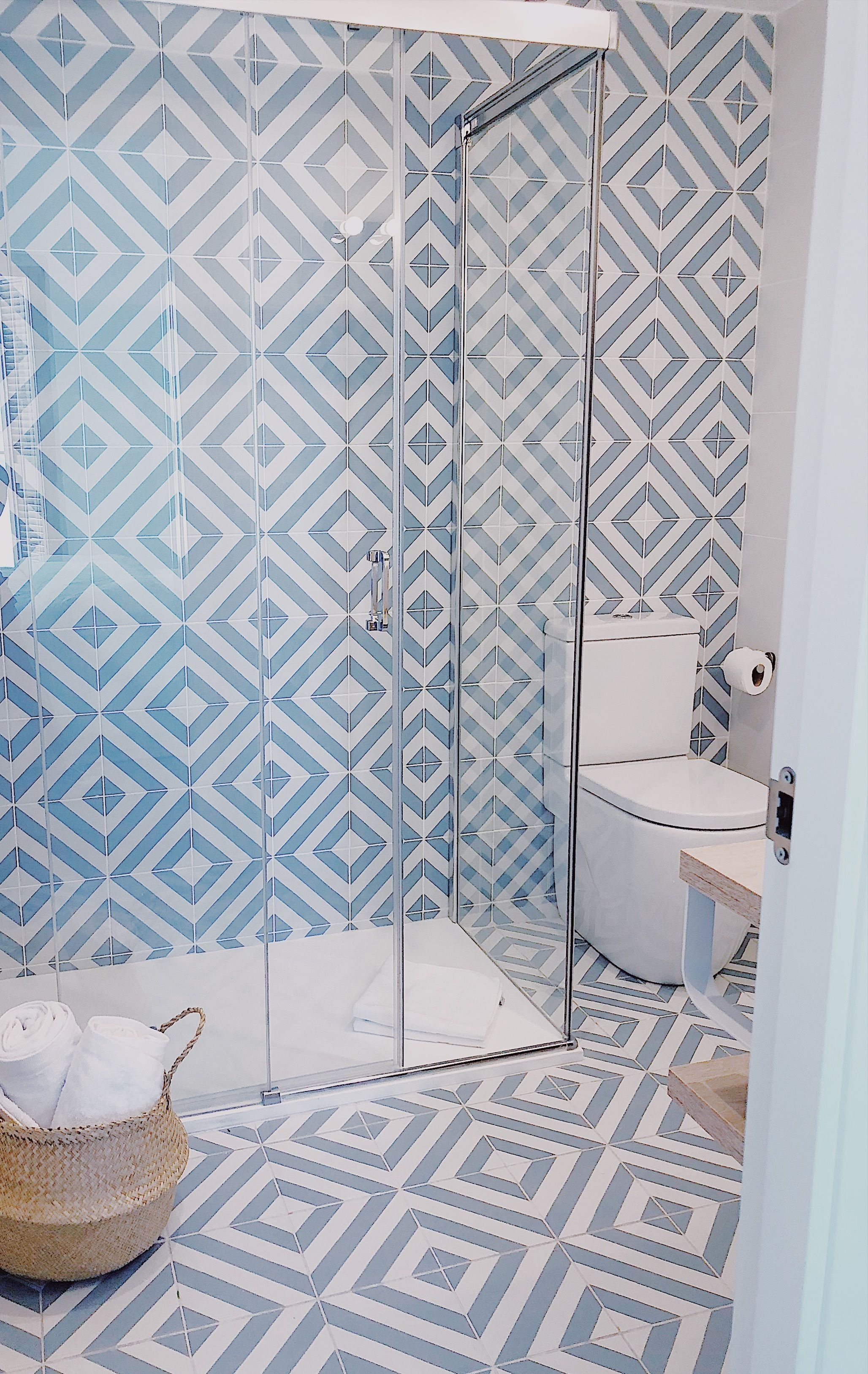 Een badkamer met een betegelde muur met een geometrisch patroon, een glazen douchecabine, een toilet en een geweven mand met handdoeken.