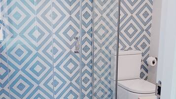 Een badkamer met een betegelde muur met een geometrisch patroon, een glazen douchecabine, een toilet en een geweven mand met handdoeken.
