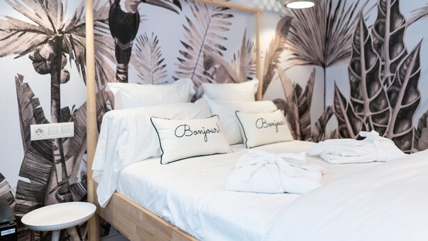 Una cama con ropa de cama blanca, dos almohadas con el texto 'Bonjour!', un cabecero de madera y un mural en la pared con hojas de palma.