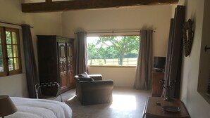 A bedroom with a bed, a sofa, a table, and a TV. There is a window with curtains and a view of trees outside. - L'auberge du Mont (Le Breuil-en-Auge)