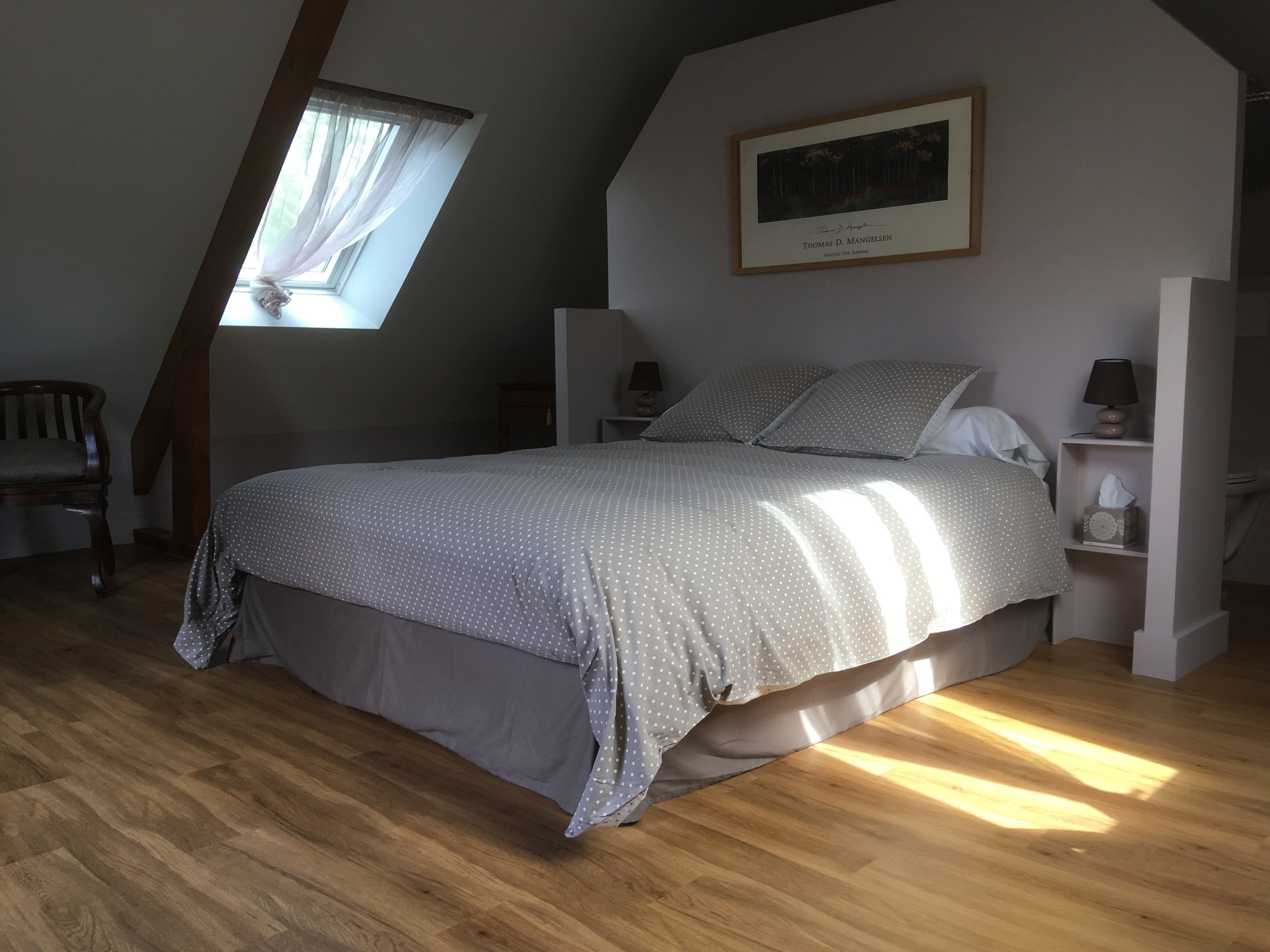 Un dormitorio con cama, mesita de noche, lámpara, silla, ventana con cortinas e imagen en un marco en la pared.