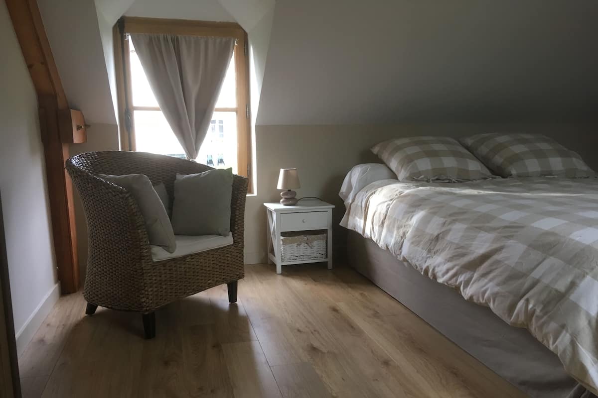 Un dormitorio con cama, una silla, una mesita de noche y una ventana con cortinas.