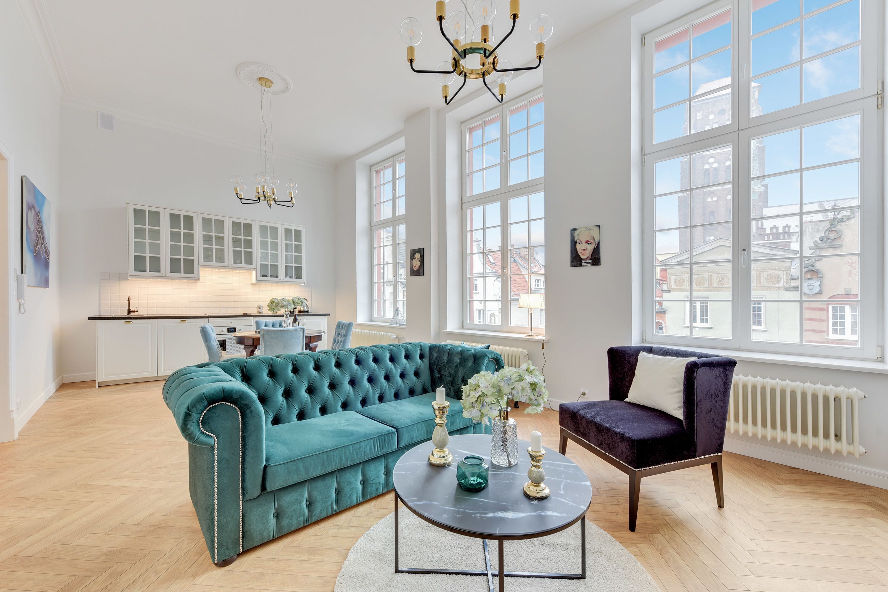 A spacious living area with a teal sofa, a round coffee table with decorative items, a dark armchair, and large windows offering a view of the city.