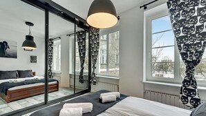 A modern bedroom with a large mirror, a bed with white linens, a dark blue headboard, and a view of the outside through a window. - Navy Group - Old Town Apartments (Gdansk)