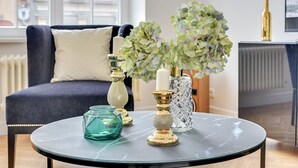 A round glass table with decorative items including a white candle, a gold candle holder, and a glass vase with green flowers. - Navy Group - Old Town Apartments (Gdansk)
