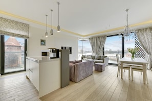 A modern living room with a kitchen area, a dining table, and a large window with a view of the city. - Navy Group - Old Town Apartments (Gdansk)