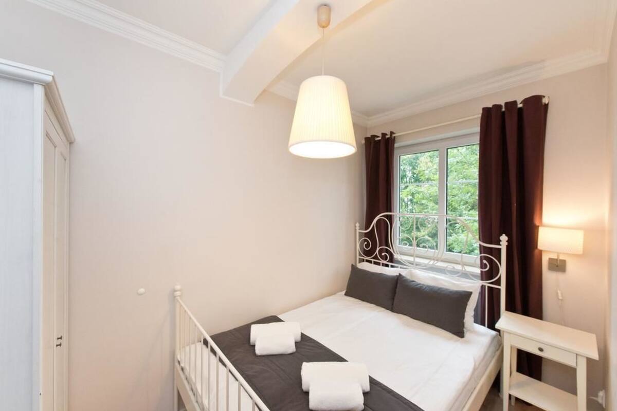 A bedroom with a bed, a bedside table, a window with curtains, and a hanging light fixture.