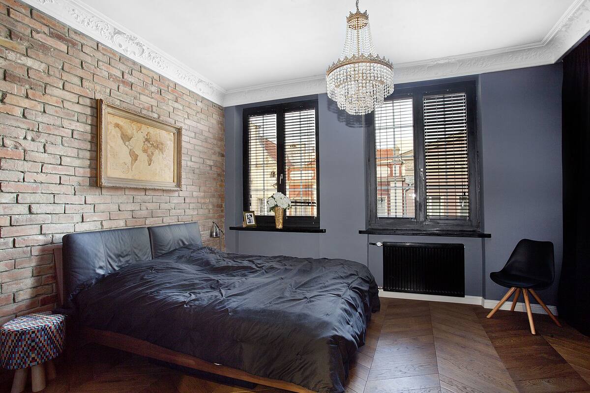 A bedroom with a brick wall, a large bed, a chandelier, and a window with blinds.