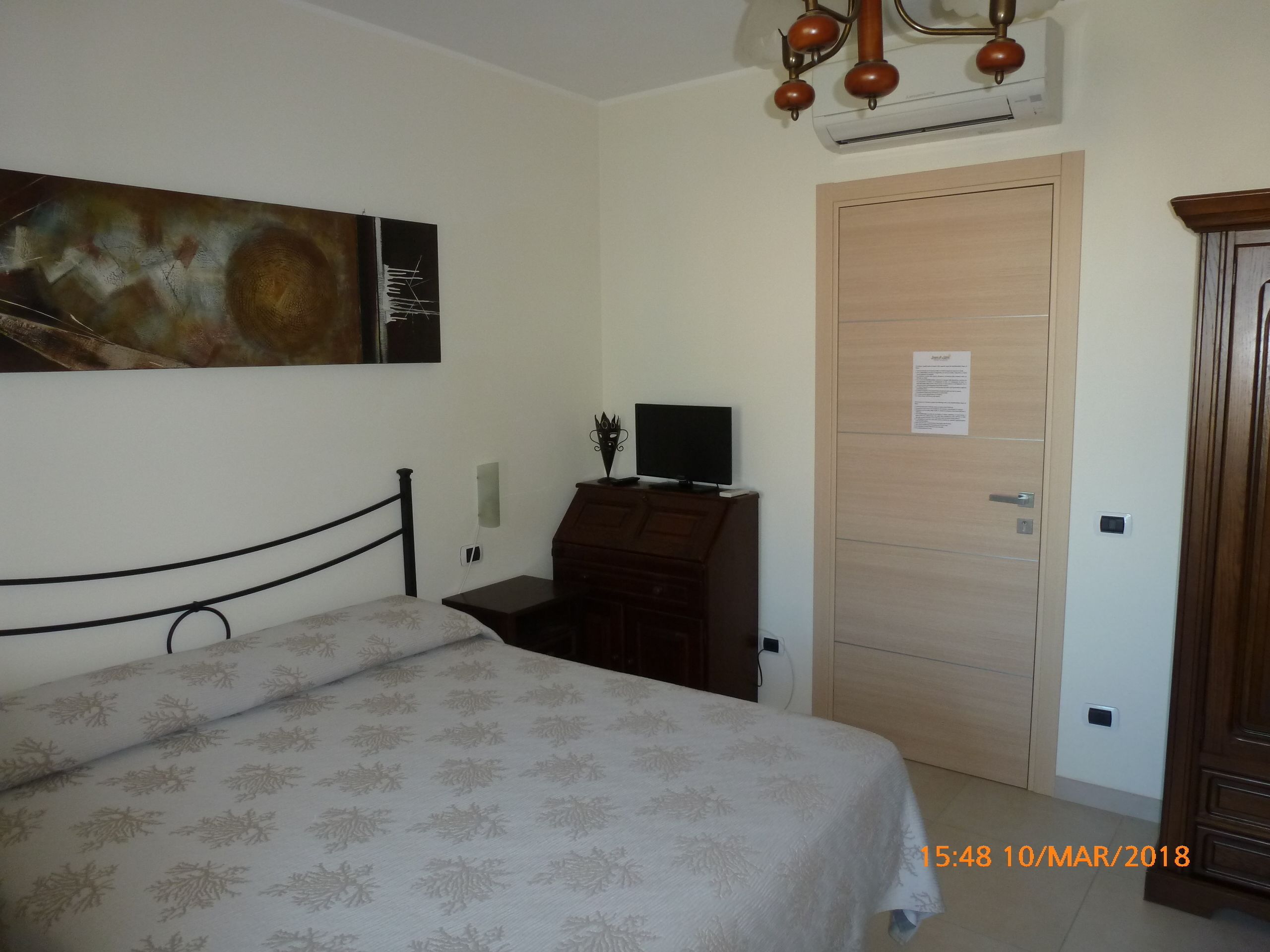 Een slaapkamer met een bed, een bureau met een computer, een houten deur en een schilderij aan de muur.