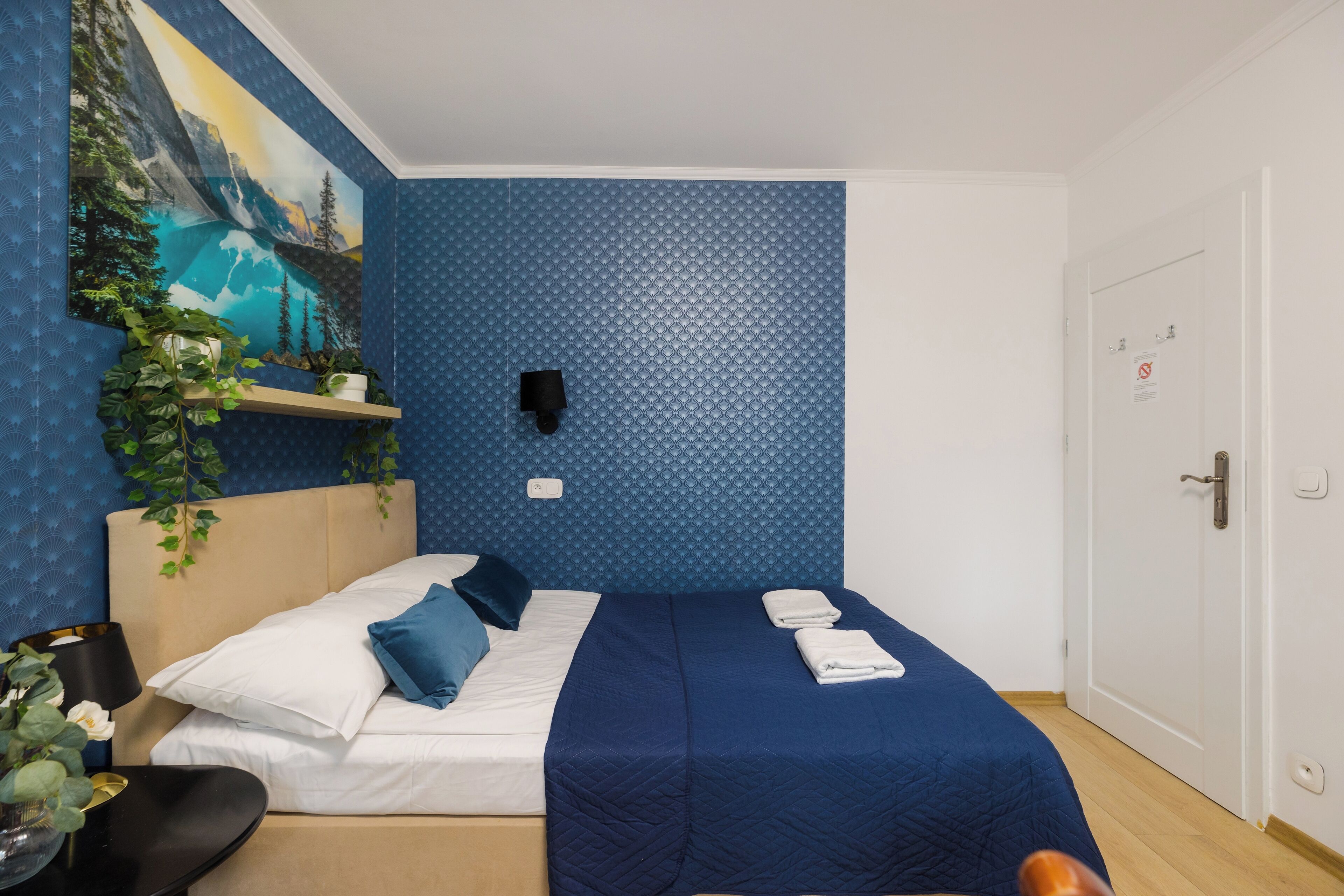 Una camera d'albergo con un letto, uno scaffale a parete con piante, una sveglia e una porta con serratura.