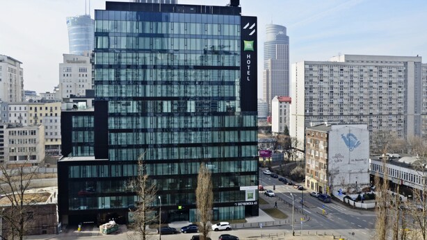 A modern hotel with a glass facade, located in an urban setting with surrounding high-rise buildings.