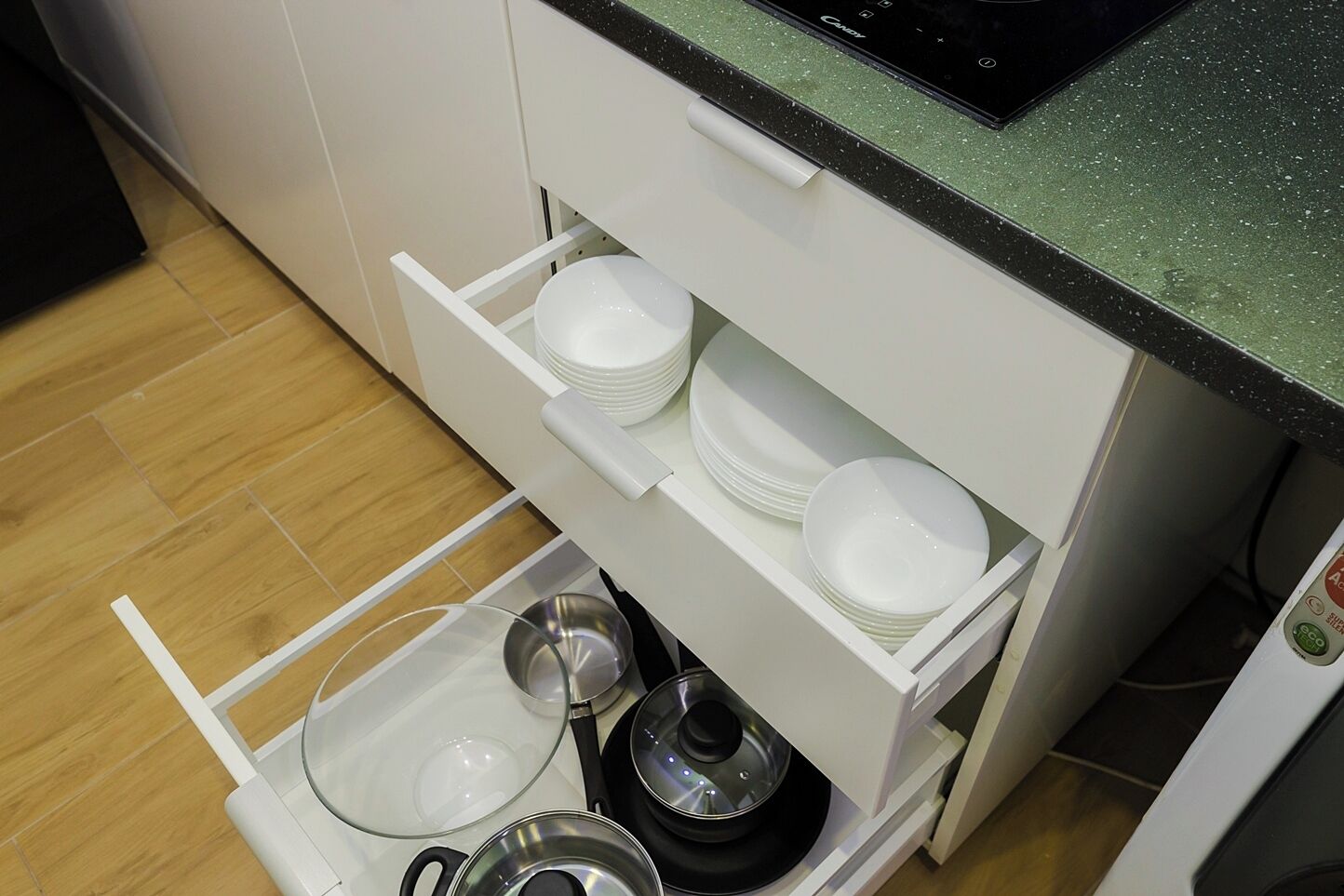 A kitchen with white cabinets, a black countertop, and a drawer with dishes and cookware.