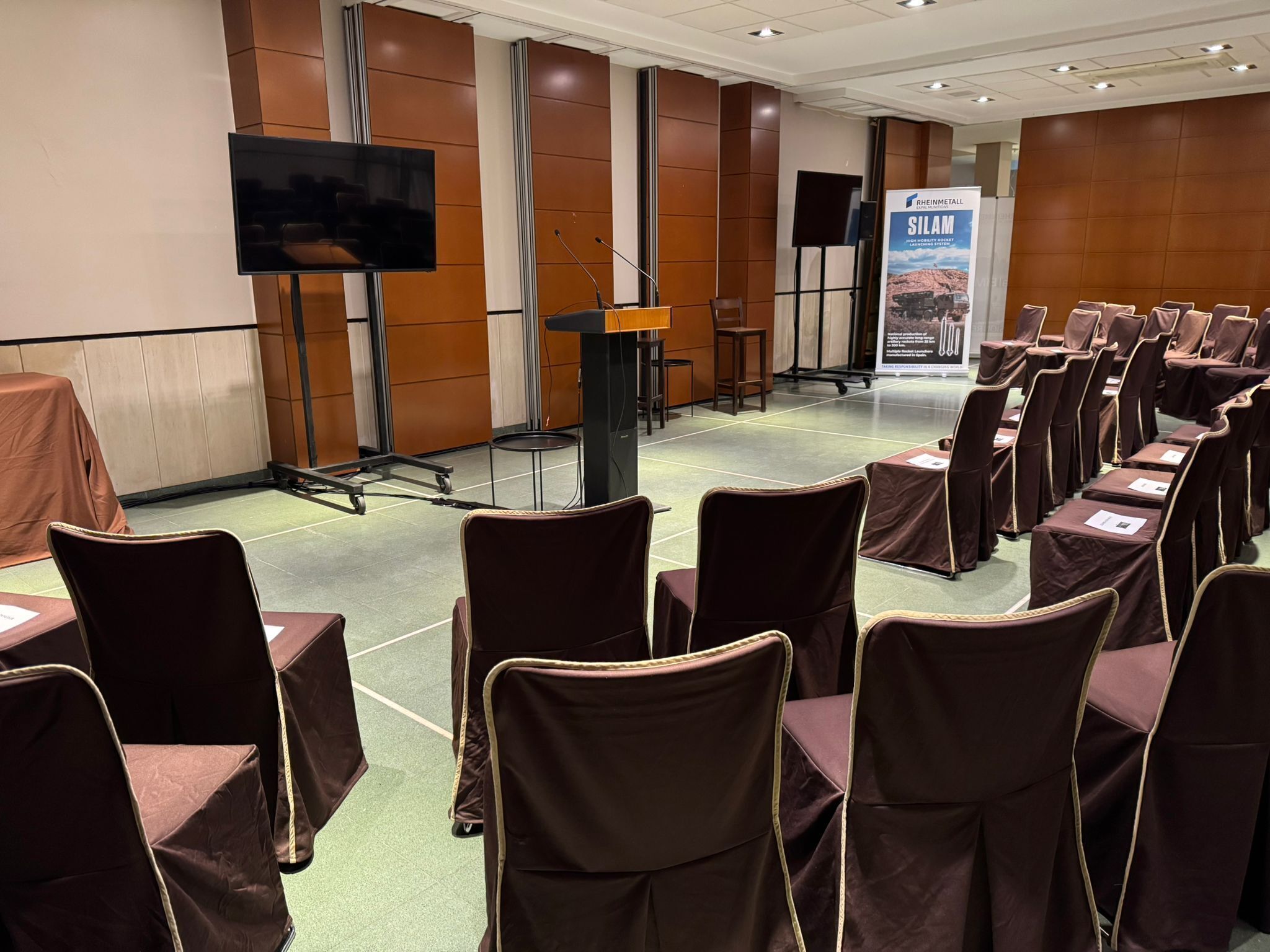 A conference room with rows of chairs, a podium, a large screen, and a poster.