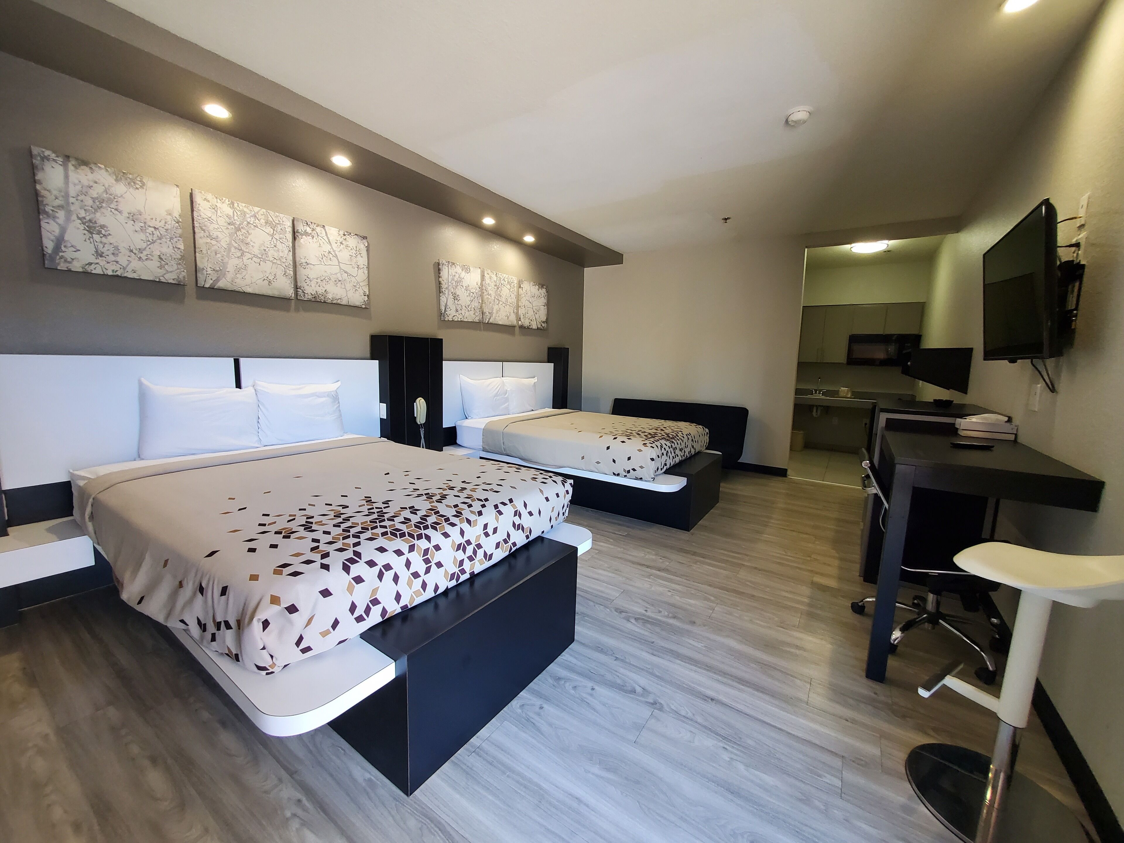 A modern hotel room with two beds, a desk, and a kitchenette.