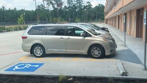 Un área de estacionamiento con señal de estacionamiento para discapacitados, una minifurgoneta color beige y un edificio con varias puertas.