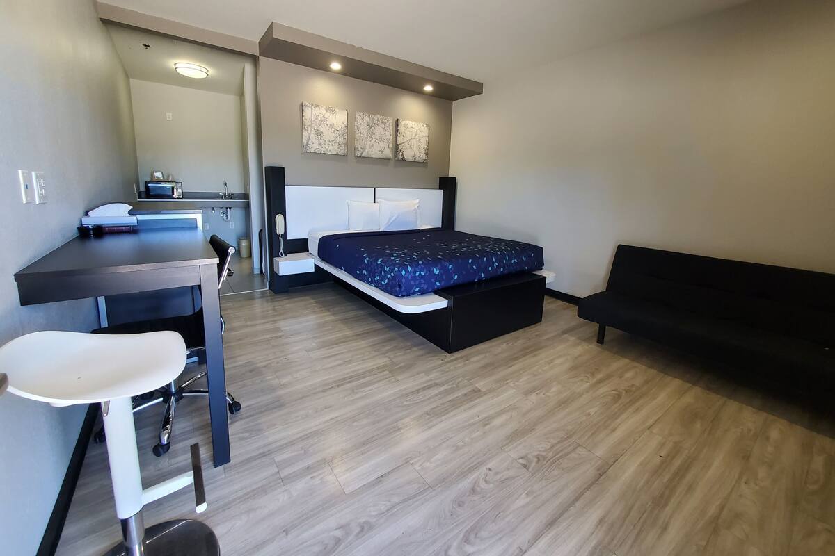 A modern bedroom with a bed, bedside table, desk, chair, and a small kitchenette area.
