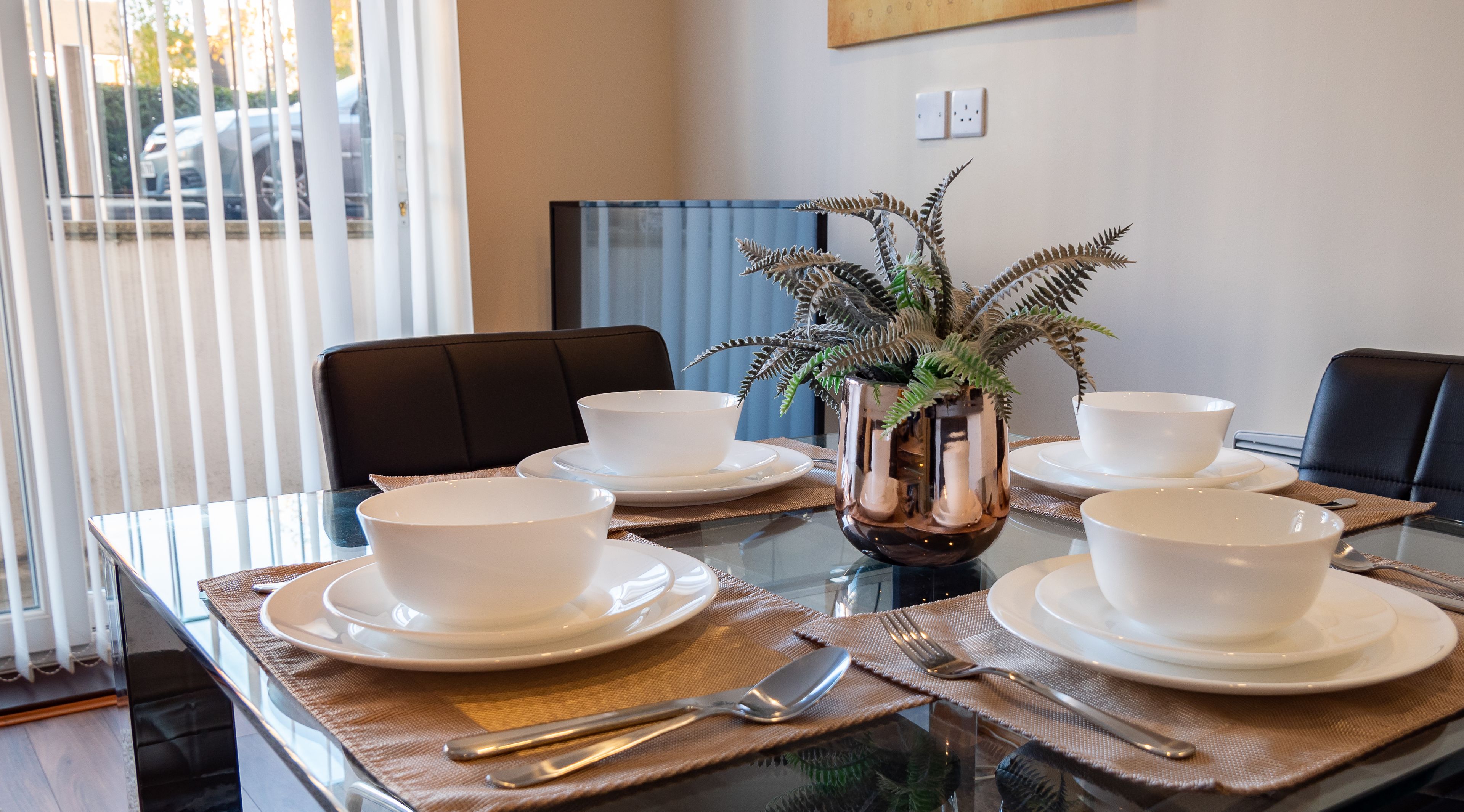 Une table à manger dressée avec des bols et des assiettes blancs, un vase contenant des feuilles vertes et une cloison en verre.