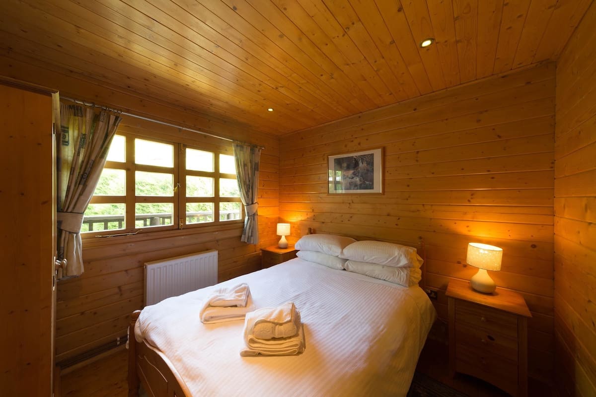 A wooden-paneled bedroom with a bed, two bedside lamps, a window with curtains, and a radiator.