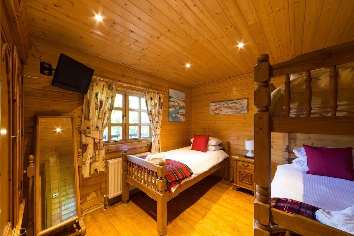 A wooden cabin room with a bed, a television, a window with curtains, and a painting on the wall.