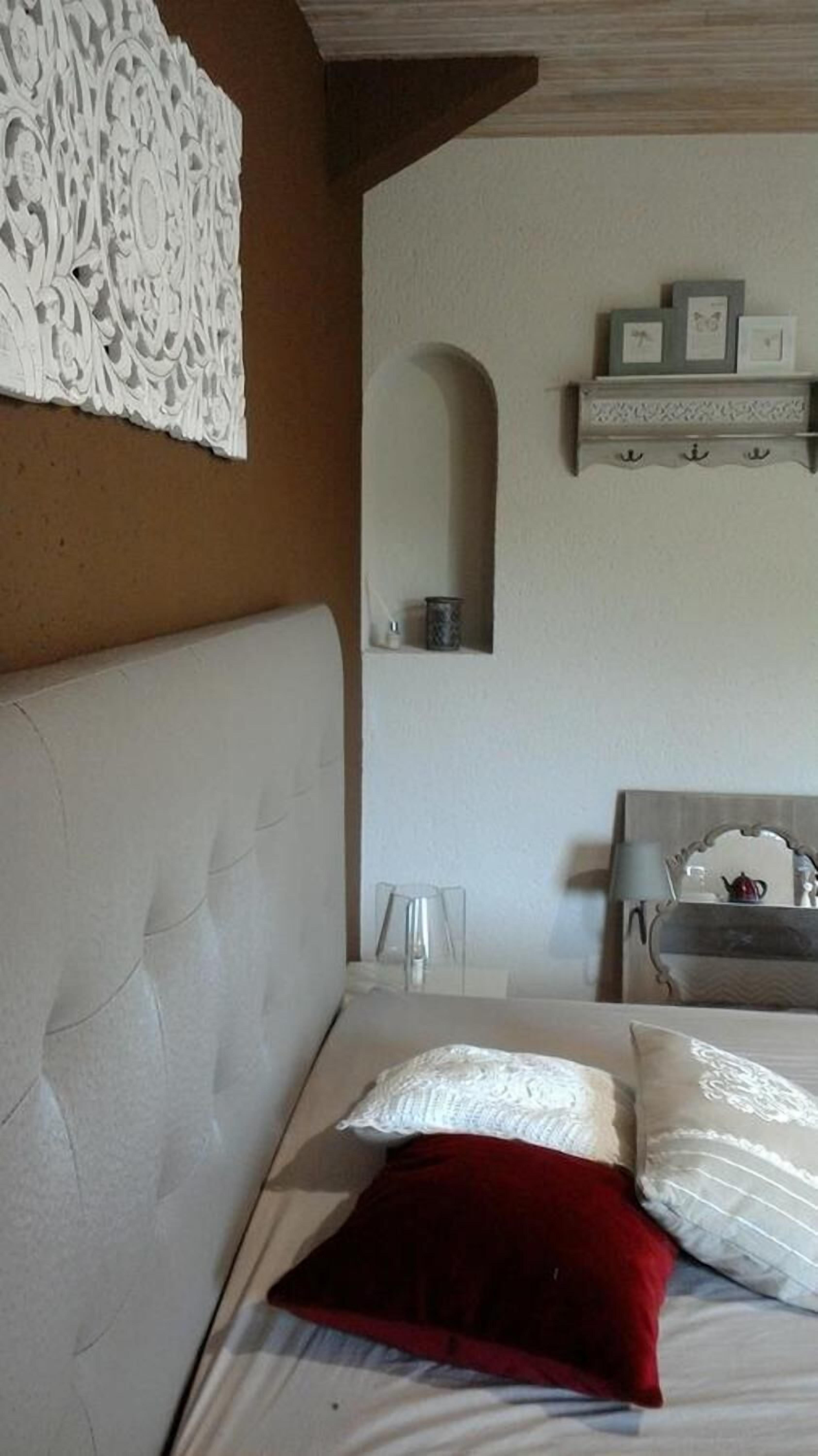 A bedroom with a tufted headboard, a bed with white and red pillows, a white wall with a decorative relief, a small alcove with a mirror, and a shelf with framed pictures.