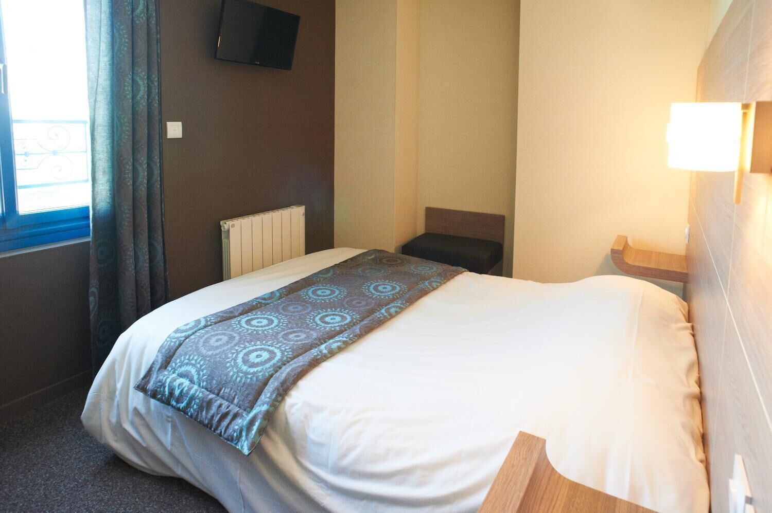 Habitación de hotel con cama, mesita de noche, lámpara, televisor en la pared y ventana con cortinas.
