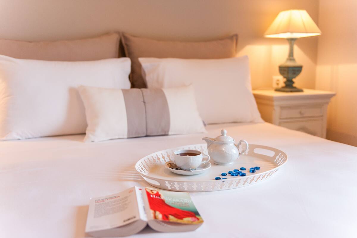 A neatly made bed with white linens, a tray with a teapot, cups, and blue candies, and a book on the bed.