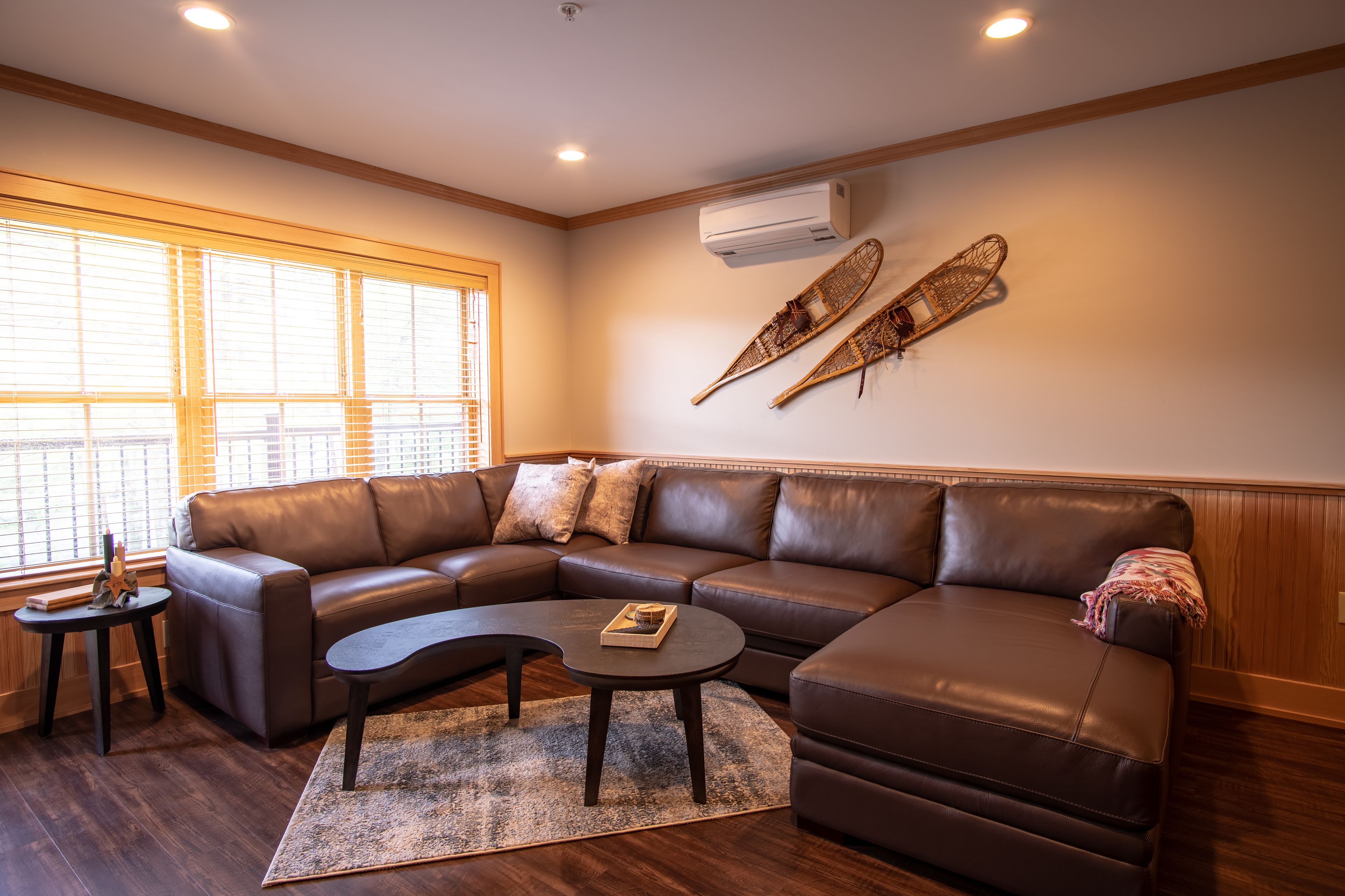 A modern living room with a brown leather sectional sofa, a round coffee table, and a small side table. There are two wall-mounted air conditioning units and two decorative paddles on the wall.