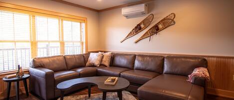 A modern living room with a brown leather sectional sofa, a round coffee table, and a small side table. There are two wall-mounted air conditioning units and two decorative paddles on the wall.