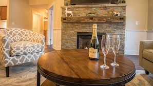A living room with a stone fireplace, a sofa, a coffee table with a bottle of champagne and two glasses, and a flat-screen TV above the fireplace. - Cranmore Condominium Lodging (North Conway)