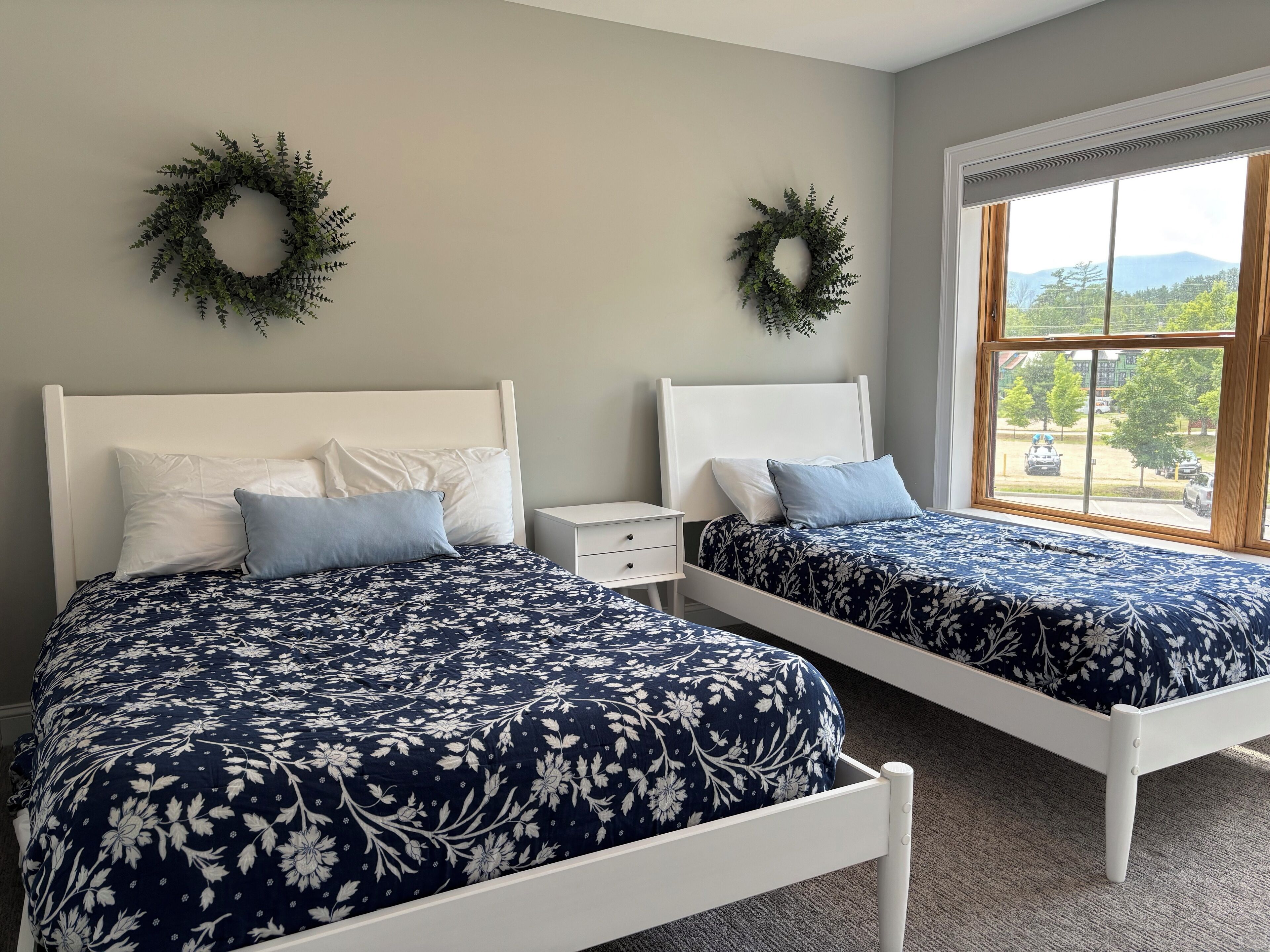 Quarto com duas camas, cada uma com colcha estampada em flor, cabeceiras brancas e almofadas combinando. Há duas mesas de cabeceira com gavetas brancas, e uma janela com vista para árvores e montanhas.