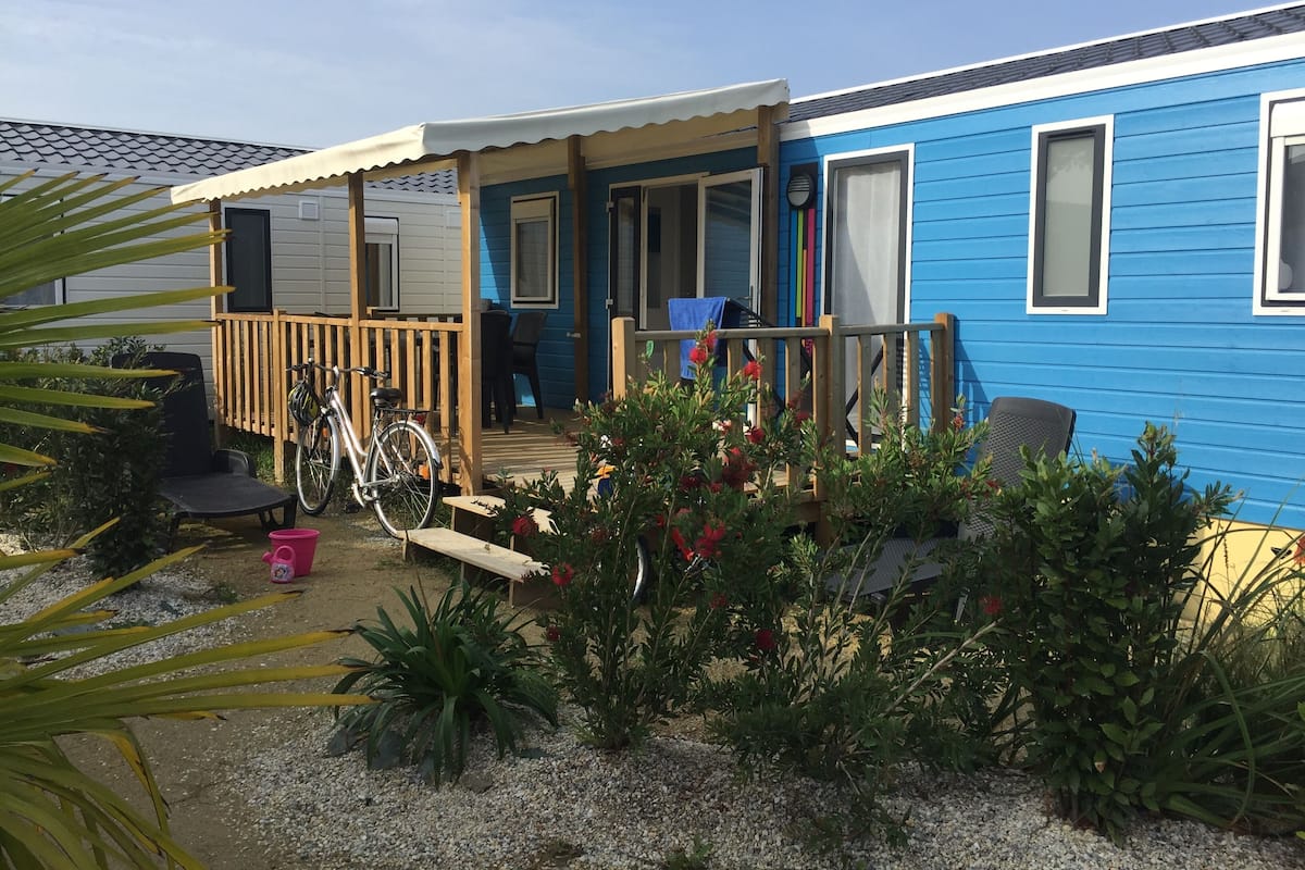 Une maison mobile bleue avec une terrasse en bois, un vélo et des meubles de jardin.