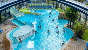 A large indoor swimming pool with multiple lanes, a slide, and a deck area with lounge chairs. - Camping Emeraude (Saint-Briac-sur-Mer)