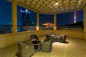 A rooftop terrace with wicker furniture and a view of city buildings at night.