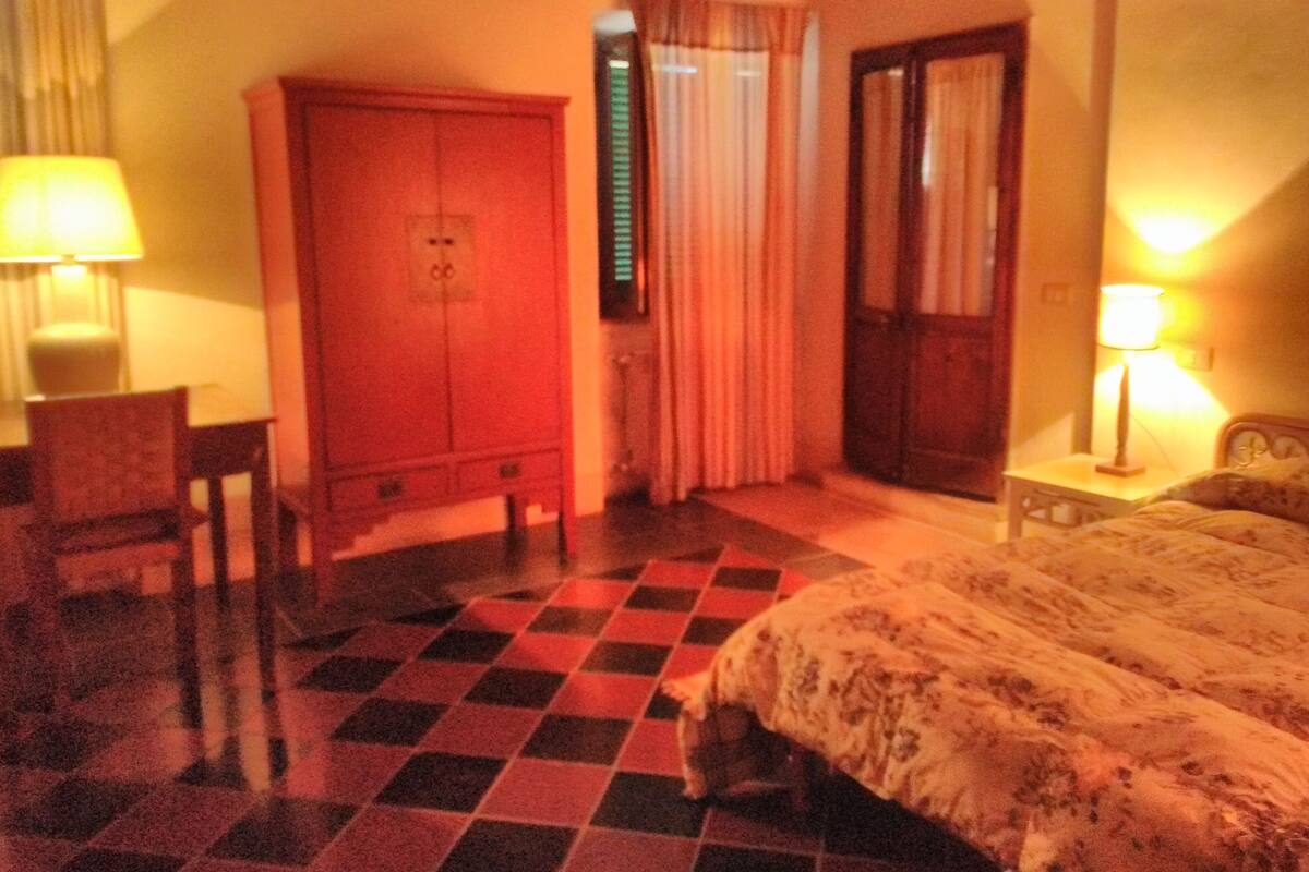 Quarto de hotel com piso xadrez, uma cama, uma escrivaninha com cadeira, um guarda-roupa e duas luminárias.