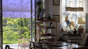 Uma zona de refeições com vista para o verde, uma mesa de madeira, cadeiras, uma estante com garrafas, um abajur e uma estátua de um burro.