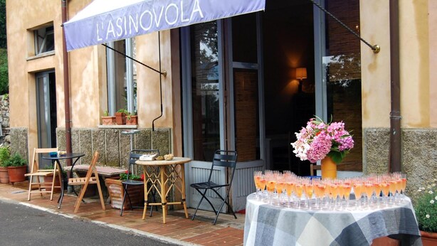 Uma área de estar ao ar livre com uma mesa posta para uma festa, um banner com a inscrição "L'ASINOVOLA" e plantas em vasos.