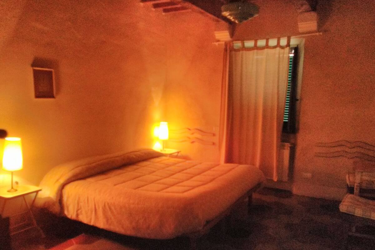 Um quarto com pouca luz, com uma cama, uma mesa de cabeceira e uma janela com cortinas.
