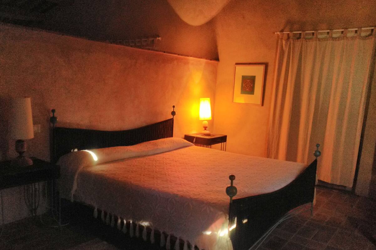 Quarto com iluminação suave, com uma cama, mesa de cabeceira com abajur e um quadro na parede.