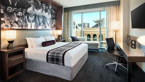 A hotel room with a large bed, a desk, a chair, a TV, and a window with a view of a building. - Texas A&M Hotel and Conference Center (College Station)